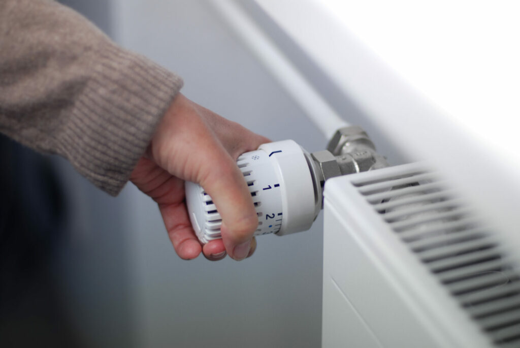 Hand dreht an einem weißen Heizungsregler, um die Raumtemperatur am Heizkörper zu regulieren.