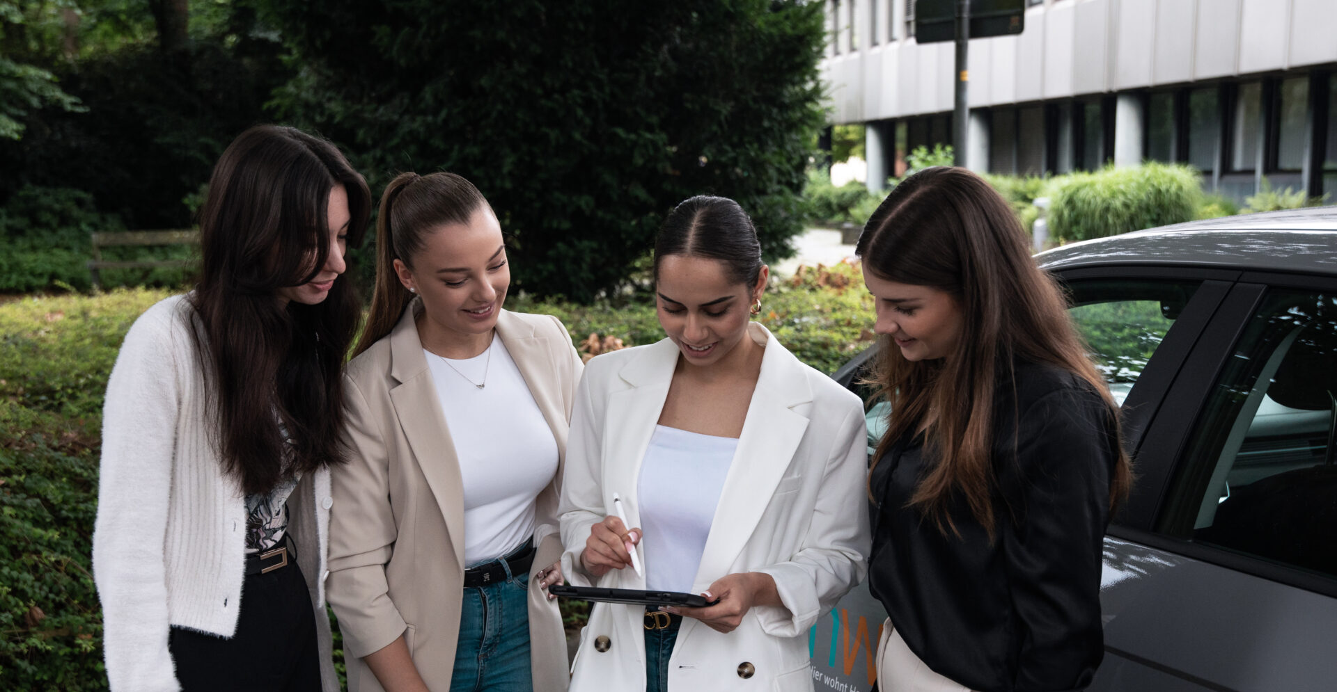Vier junge Frauen stehen vor einem Firmenwagen der HWG und schauen gemeinsam auf ein Tablet. Eine Frau in weißem Blazer hält das Gerät und schreibt mit einem digitalen Stift. Die Szene spielt im Grünen vor einem Bürogebäude und wirkt modern, kooperativ und professionell.